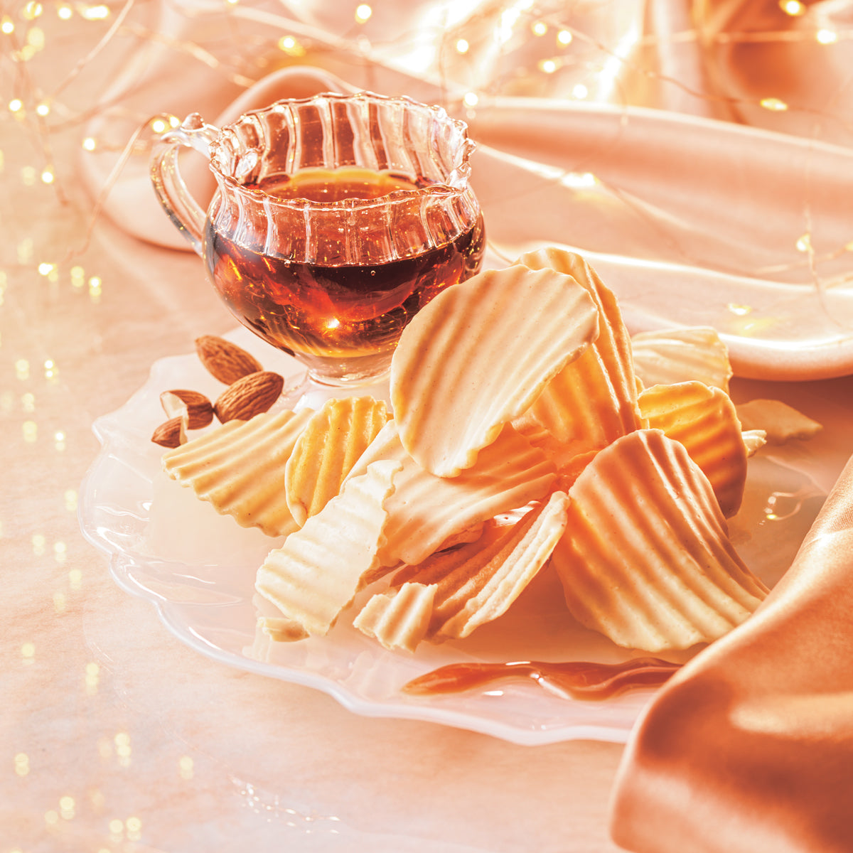 Potatochip Chocolate "Maple Nutty" - Image shows golden, ridged chocolate-coated potato chips on a clear plate with almonds and a cup with maple syrup. Accents include golden lights and a peach-colored tablecloth. Surface is in light peach.